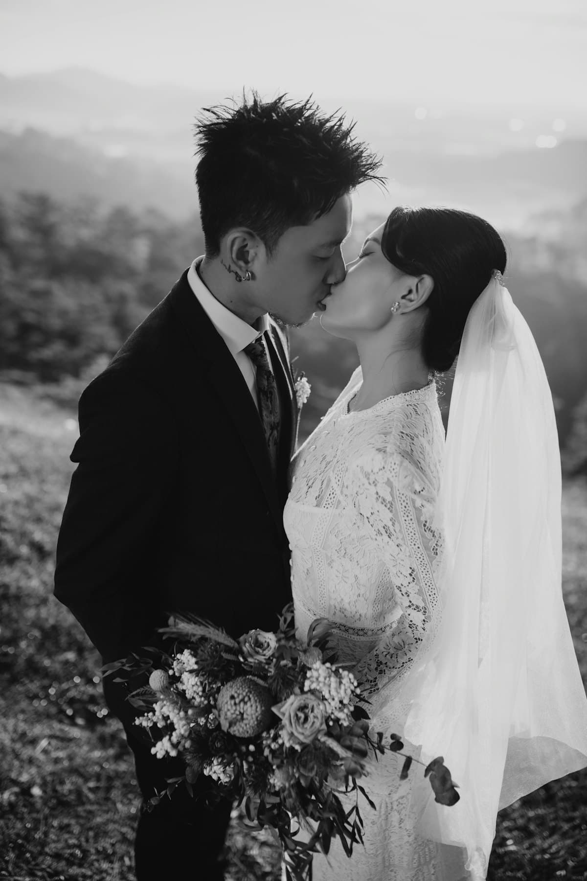 Cô dâu cầm bó hoa và chú rể hôn nhau trên sườn đồi, phía sau là núi trong sương